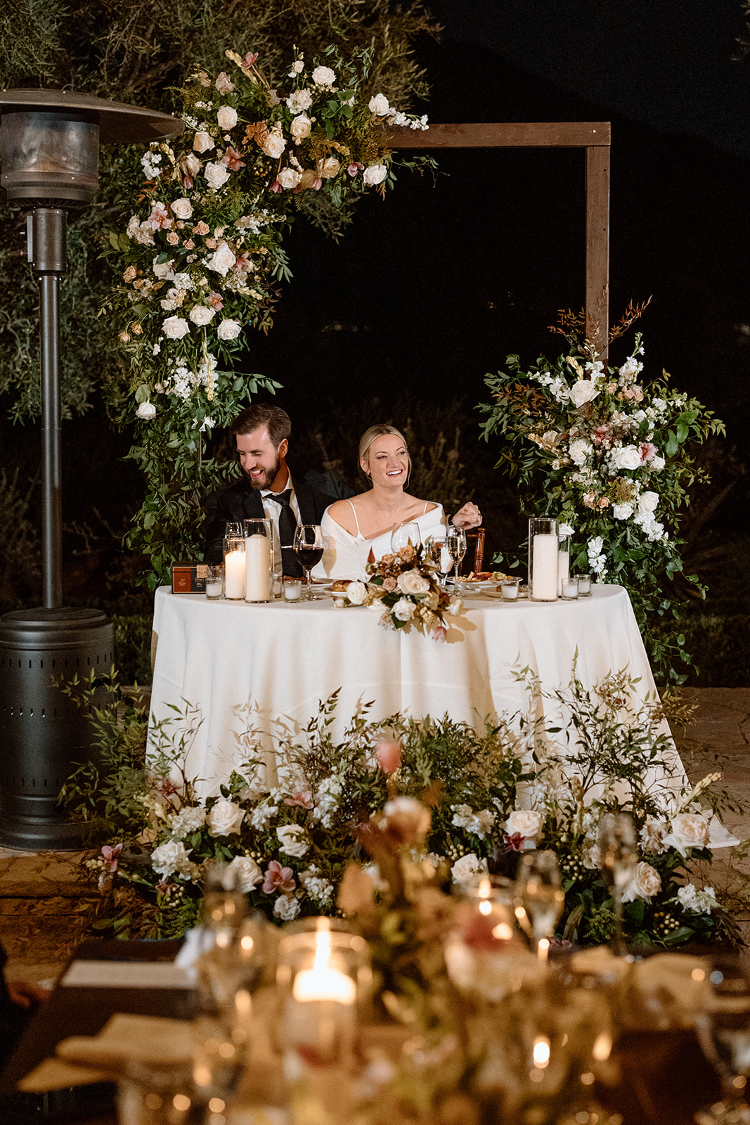 Bride and groom sit at their reception table while guests give speeches at their wedding